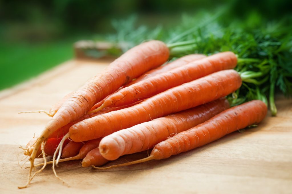 a bunch of carrots, unpeeled and with their stems in tact.  They are laying down on a cutting board. 